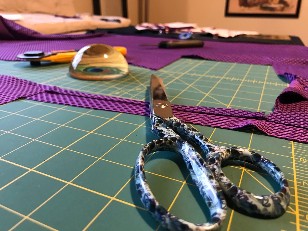 Photo of a sewing table, close up on a pair of scissors, with fabric and other fabric-cutting tools in the distance. 